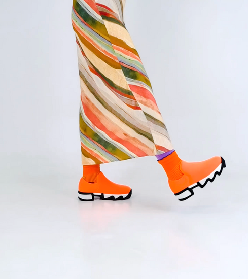 A model walking and twirling in WES Neon Orange Low Top Sneaker and DOUBLE DECK Carrot Crew Sock. Styled with shoulder strapped dress with multi colored print.