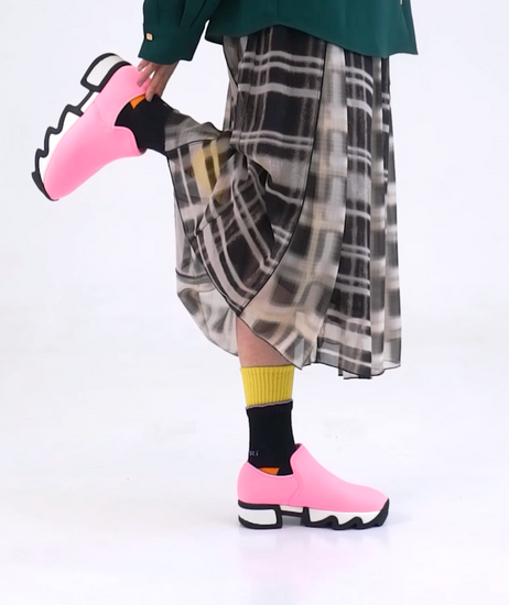 A model wearing WES Pink Low Top Sneaker with DOUBLE DECK Black Multi socks, walking and dancing. Styled with checked flared skirt and deep green shirt.